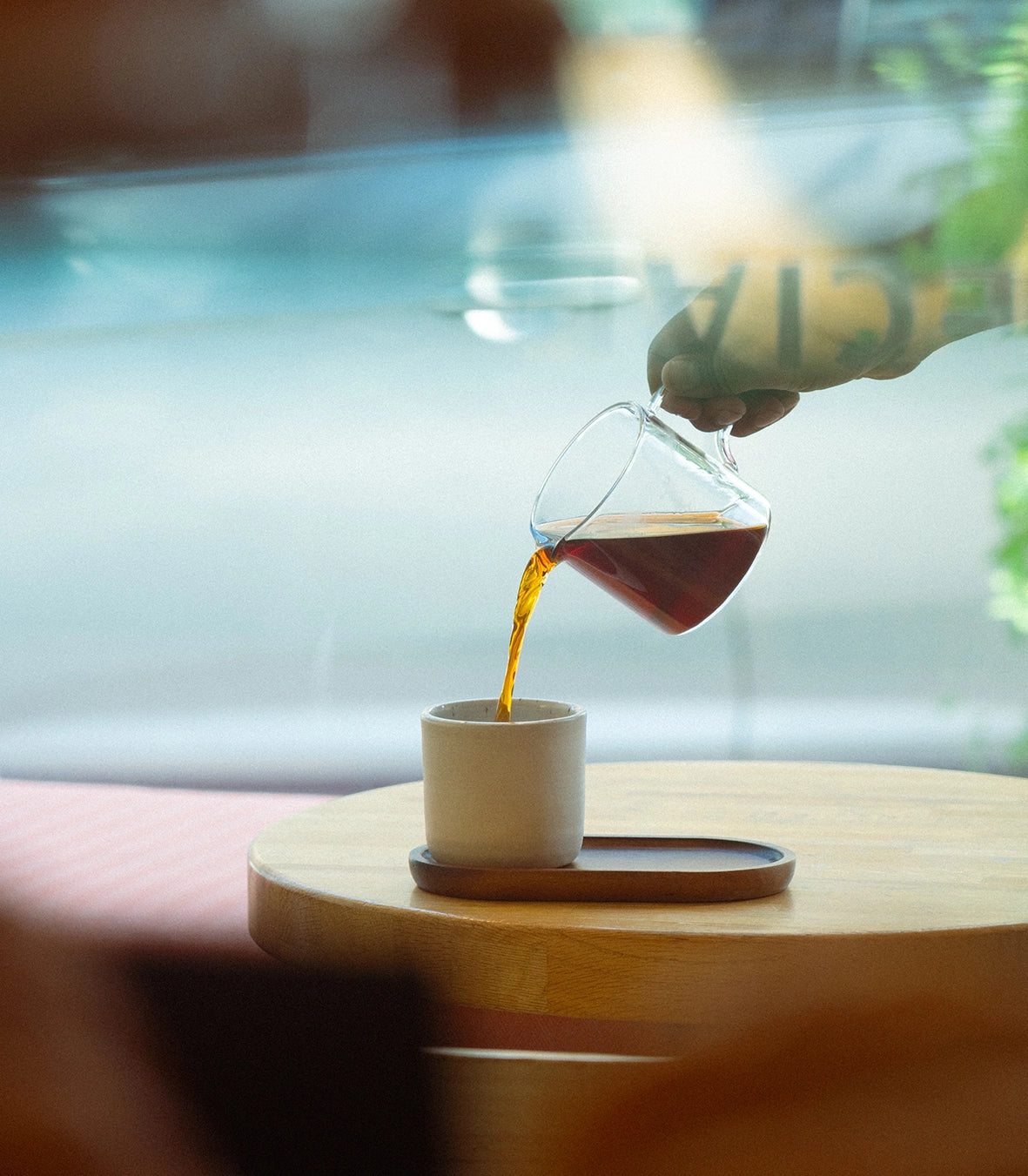 A person pouring filter coffee from a pot into a ceramic cup that is placed on a wooden plate on a table.